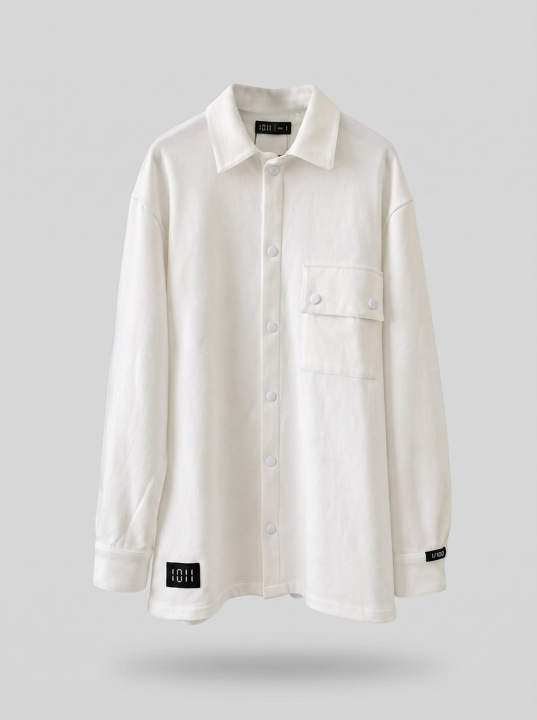 An off-white long-sleeve button-up shirt with a collar, a chest pocket, and small black brand patches near the cuffs, shown against a solid gray background.